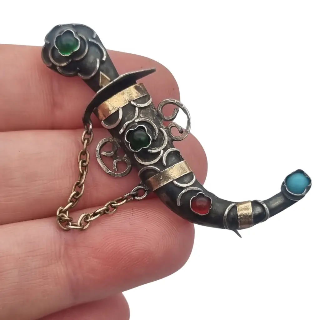 Broche de plata en forma daga con detalles dorados y pedrería colores